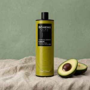 Flacon de shampooing kératine et avocat Bôheme Secret Paris posé sur du lin naturel devant un fond vert sauge, avec un avocat coupé en deux